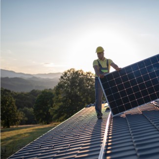 A worker in a yellow hard hat and reflective vest is installing a large solar panel on a sloped rooftop. The background features a scenic view of rolling hills and trees under a soft sunset sky, highlighting the use of renewable energy in a natural setting.