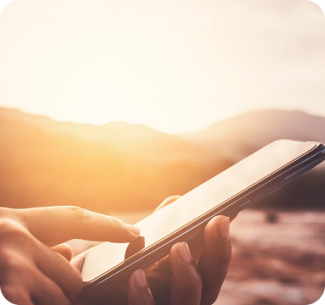Close-up of a person’s hand using a smartphone outdoors with a bright sunset and mountains in the background.